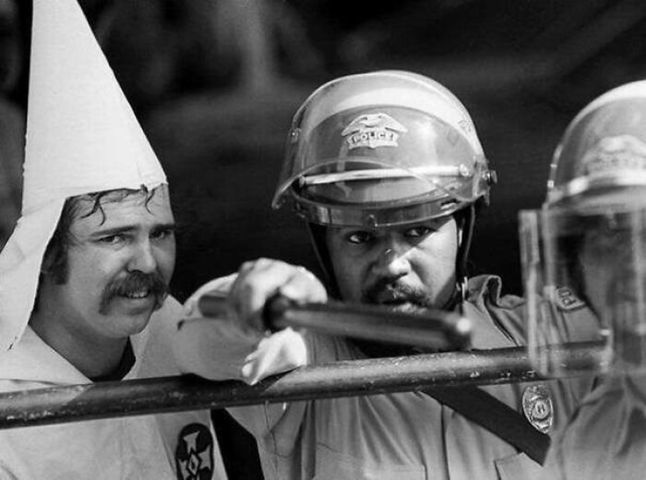 Członek Ku Klux Klanu okrywający się za policjantem po tym, jak ludzie otoczyli pochód w Austin w Teksasie (1983).