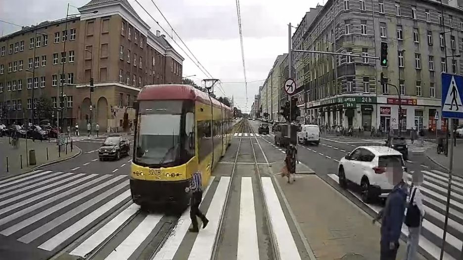 Tramwaj w żółto-czerwonym kolorze przejeżdża przez przejście dla pieszych, obok ruchliwej ulicy z samochodami i pieszymi.