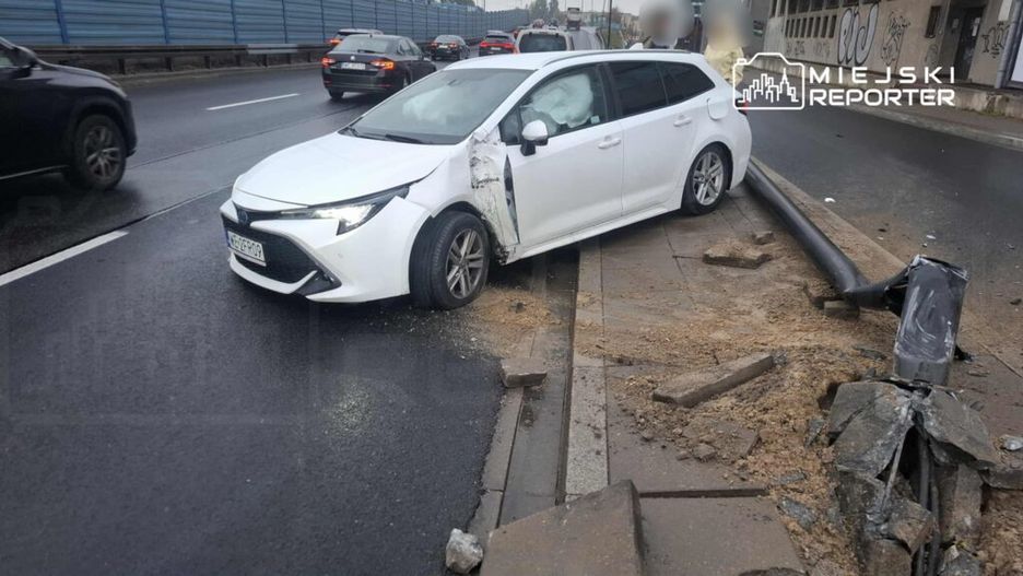 Biały samochód osobowy stoi na poboczu drogi, z uszkodzoną lewą stroną i otwartymi drzwiami. W tle widać inne pojazdy na jezdni.