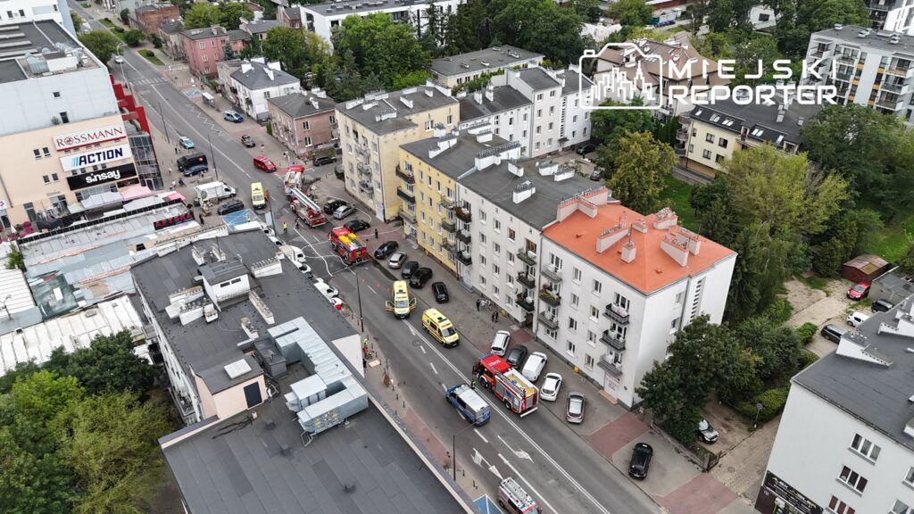 Na ulicy widać wiele pojazdów ratunkowych, w tym samochody straży pożarnej i karetki, otaczających budynek mieszkalny.