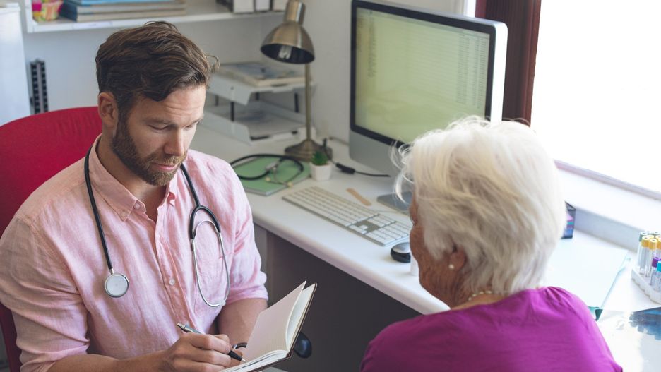 Front view of a confident Caucasian male doctor writing prescription on notebook for senior woman in clinic room