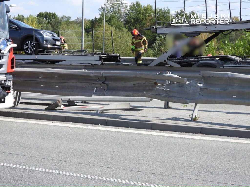 Uszkodzona bariera ochronna na drodze, obok stojący wóz strażacki oraz strażacy w akcji.