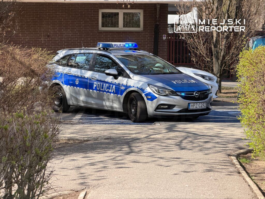 Policyjny samochód osobowy z niebieskimi światłami zaparkowany na chodniku w otoczeniu drzew i krzewów.