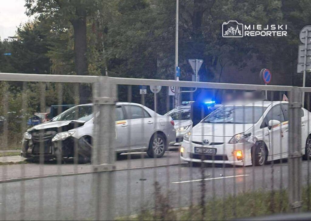 Dwa uszkodzone samochody osobowe stoją na poboczu, a obok nich policyjny radiowóz z włączonymi sygnałami świetlnymi.
