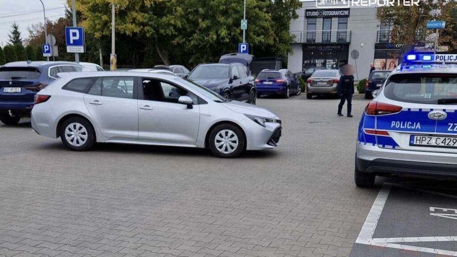 Srebrny samochód osobowy manewrujący na parkingu, obok radiowóz policyjny z włączonymi sygnałami świetlnymi.