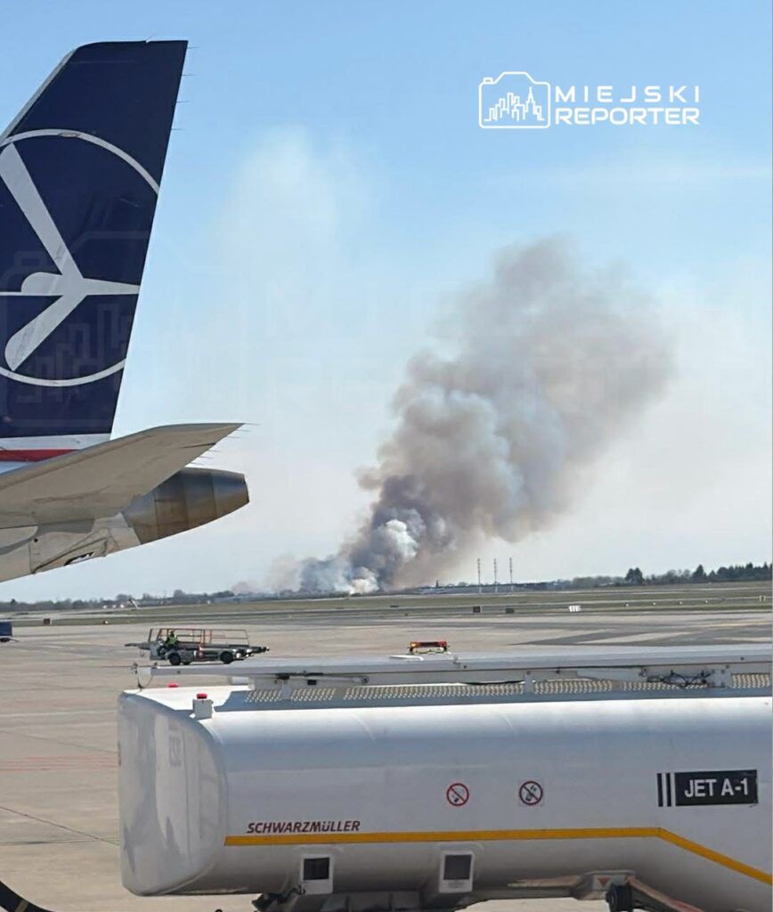 Samolot z logo linii lotniczej na pierwszym planie, w tle widoczny dym unoszący się z pożaru na horyzoncie.