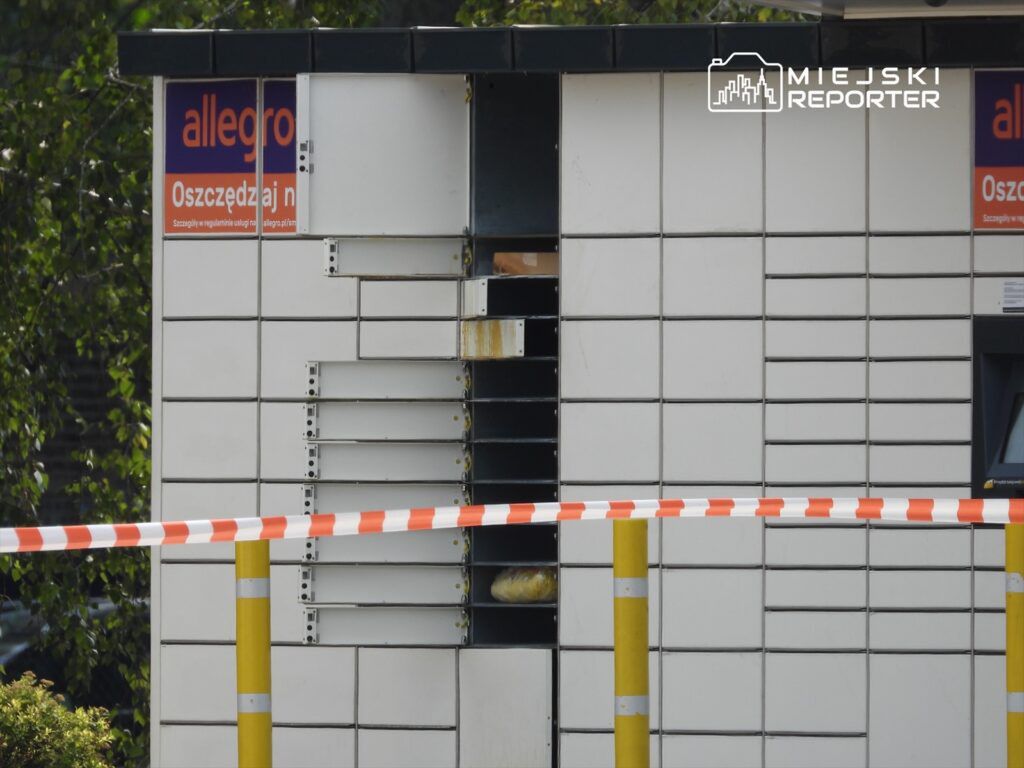 Uszkodzony paczkomat z otwartymi drzwiczkami, obok bariera w biało-czerwone paski, w tle zieleń drzew.