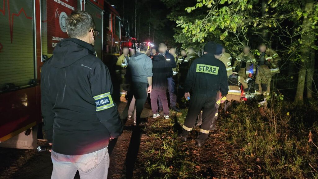 Grupa ratowników i policjantów z latarkami prowadzi akcję w lesie w nocy, obok wozu strażackiego.