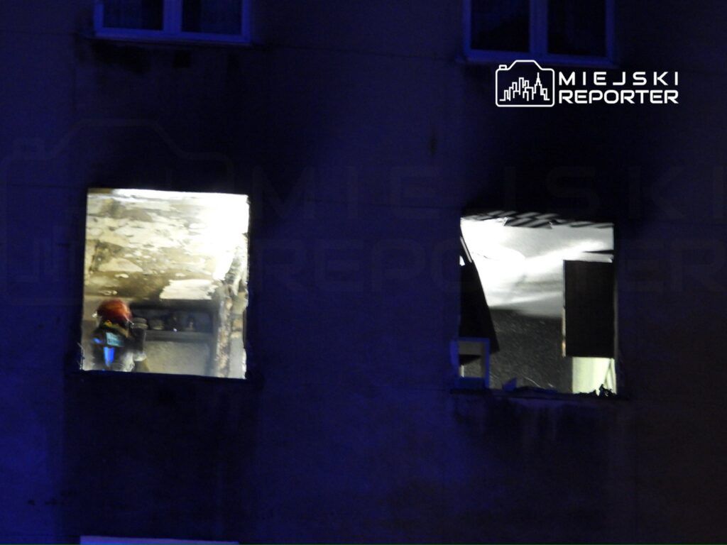 Strażak w mundurze z aparatem tlenowym przeszukuje zniszczoną kuchnię przez okno budynku.