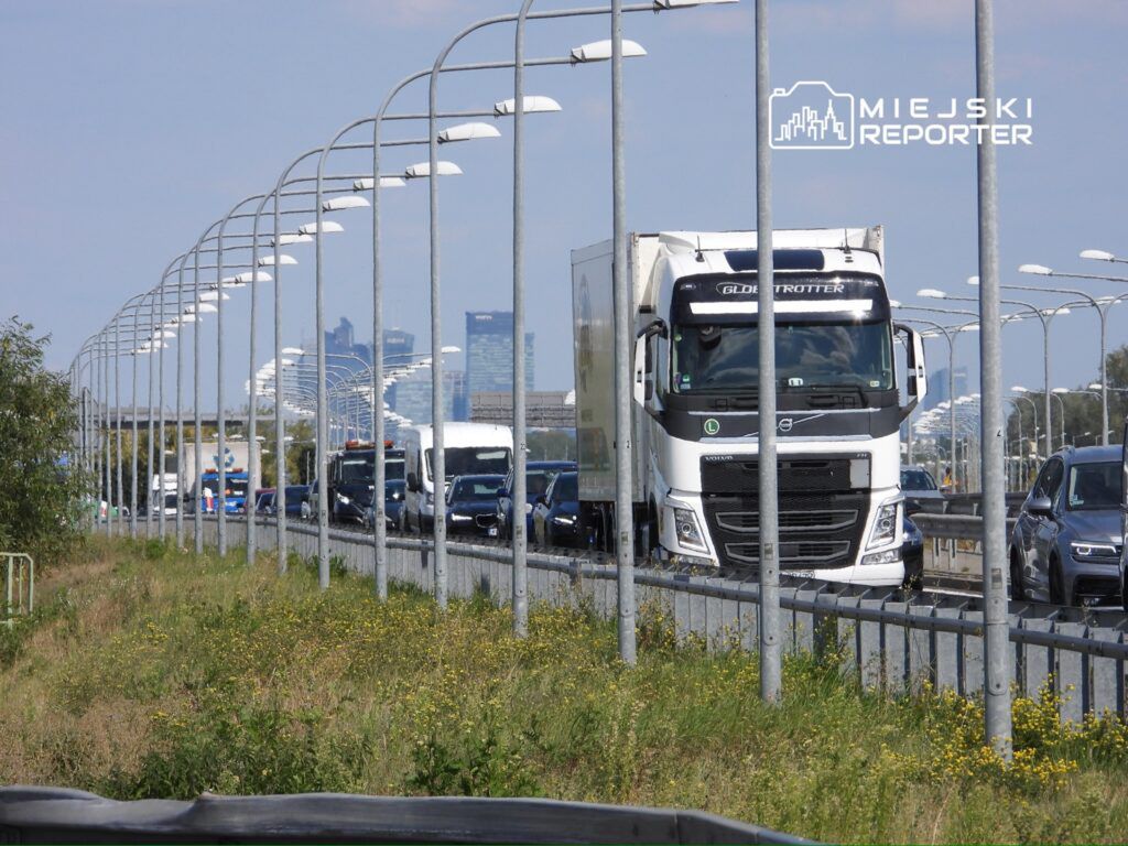 Ruch samochodowy na drodze ekspresowej z widocznym ciężarowym pojazdem marki Volvo oraz nowoczesnym miastem w tle.