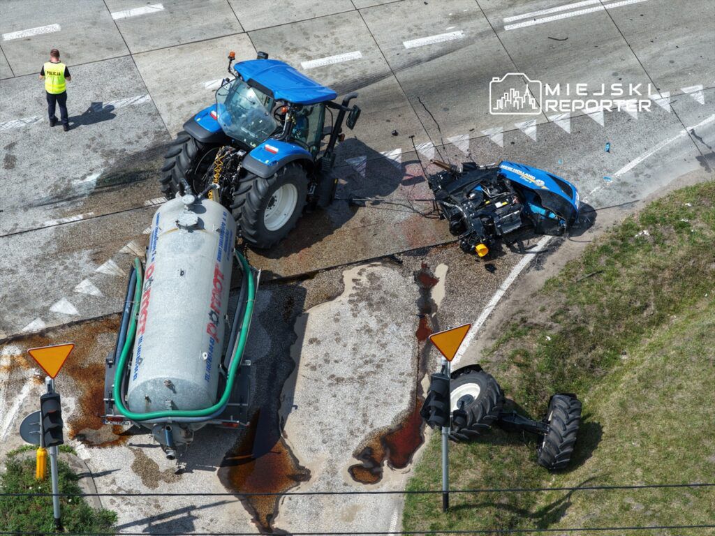 Traktor z uszkodzoną przyczepą stoi na drodze, obok policjant kontrolujący sytuację. Na jezdni widoczne plamy cieczy.