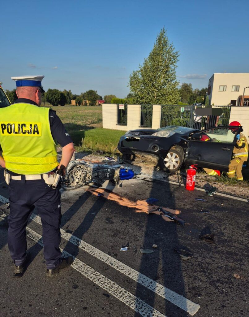 Policjant w kamizelce odblaskowej obserwuje wypadek drogowy z uszkodzonym samochodem i wrakiem motocykla.