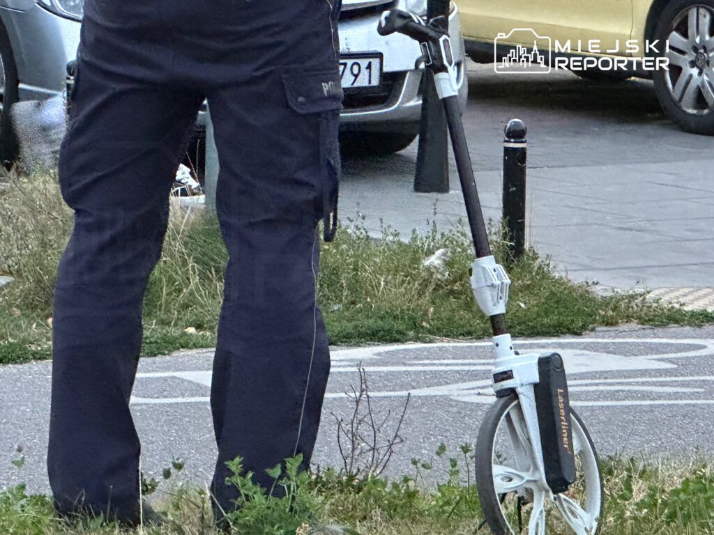 Mężczyzna w granatowych spodniach policyjnych stoi obok zaparkowanej hulajnogi na chodniku.