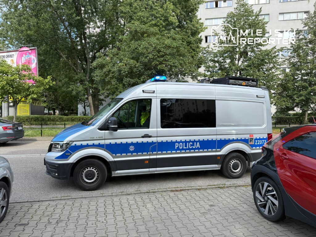 Policyjny radiowóz z niebieskimi światłami zaparkowany obok chodnika w otoczeniu drzew i budynków mieszkalnych.