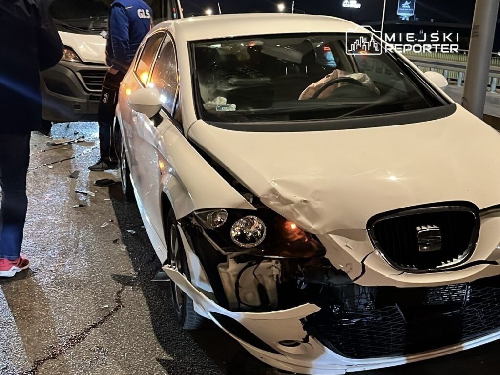 Biały samochód osobowy z uszkodzoną przednią częścią stoi na jezdni, obok dwóch mężczyzn rozmawiających w nocy.