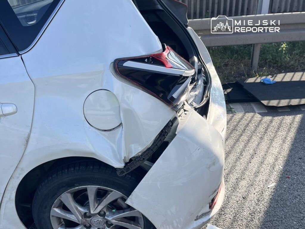 Uszkodzone białe auto osobowe z wgniecioną tylną częścią na poboczu drogi, obok leży fragment zderzaka.
