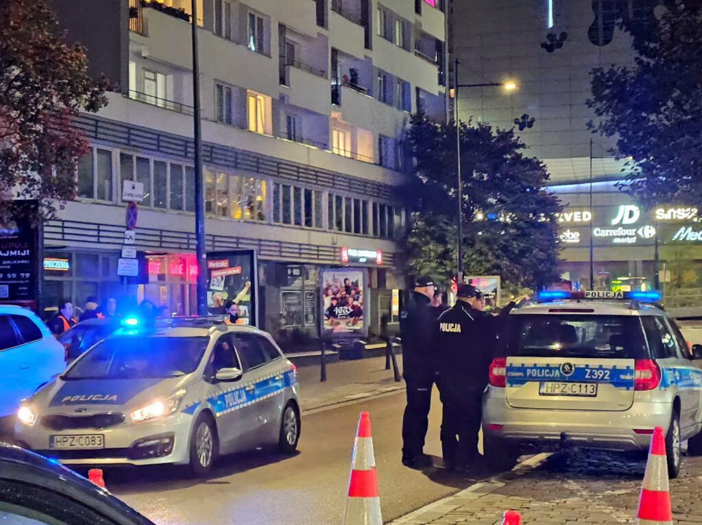 Grupa policjantów rozmawia obok radiowozu z włączonymi sygnałami świetlnymi na oświetlonej ulicy.