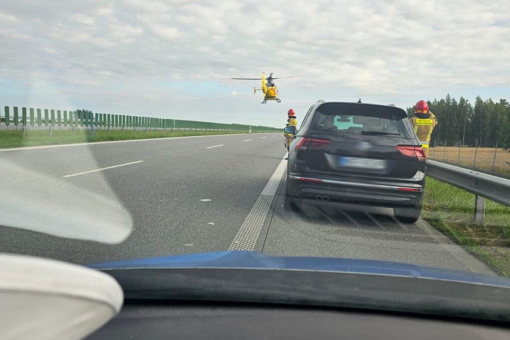 Śmigłowiec ratunkowy przelatuje nad autostradą, podczas gdy strażacy stoją obok zatrzymanego samochodu.