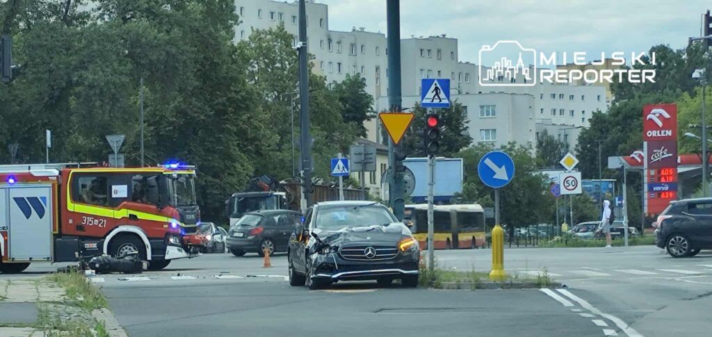 Na zdjęciu widoczny jest wypadek drogowy, w którym uczestniczy czarny samochód marki Mercedes z uszkodzoną przednią częścią. Obok znajduje się wóz strażacki z włączonymi sygnałami świetlnymi. W tle widać inne pojazdy oraz znaki drogowe. Zdarzenie miało miejsce na ruchliwej ulicy.