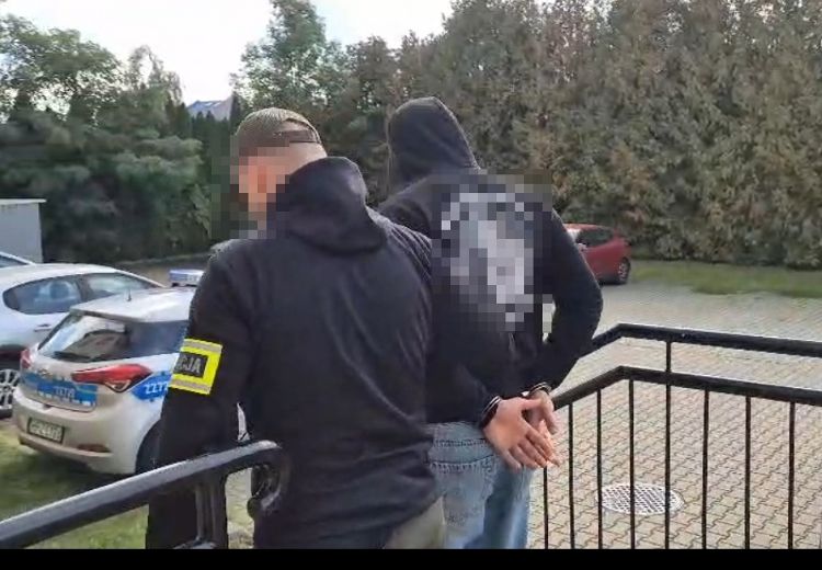 Mężczyzna w ciemnym ubraniu jest prowadzony przez policjanta na zewnątrz budynku, w tle widać radiowóz.