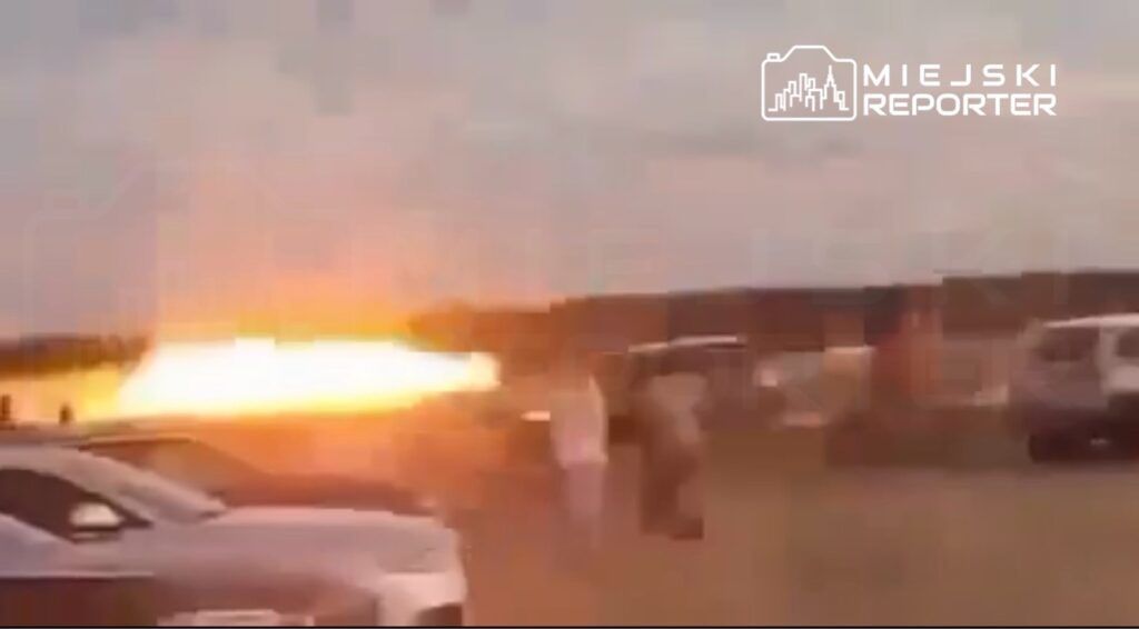 Grupa ludzi obserwująca wybuchający ogień na tle zachodzącego słońca, w pobliżu zaparkowanych samochodów.