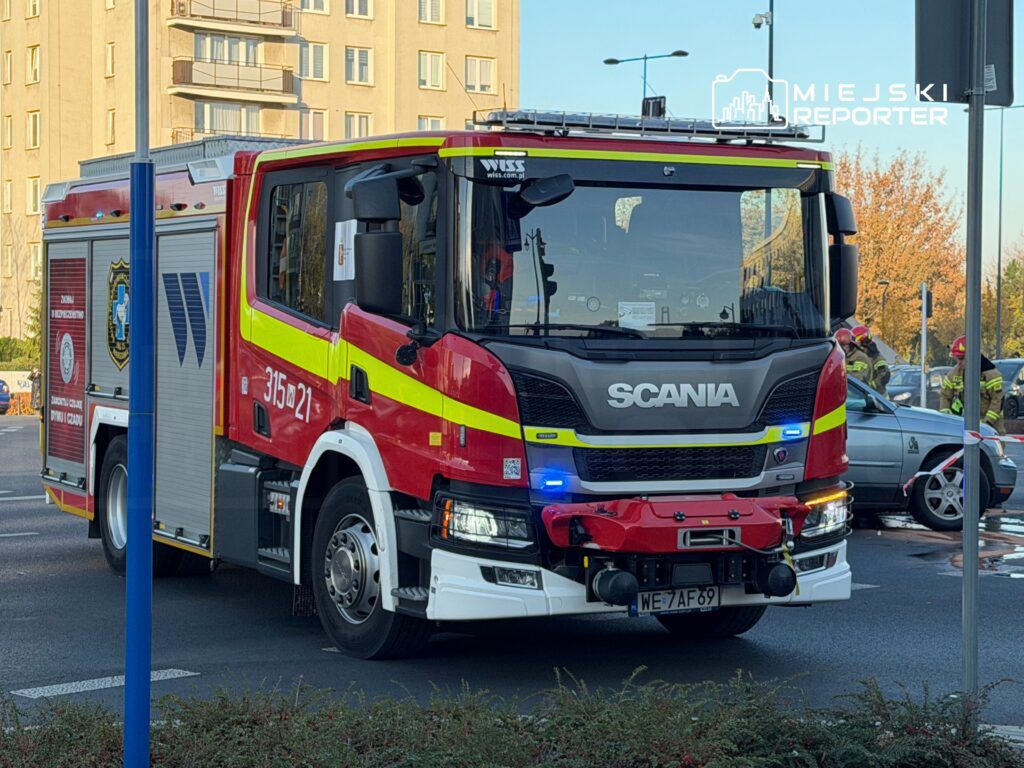 Czerwony wóz strażacki marki Scania z włączonymi sygnałami świetlnymi stoi na skrzyżowaniu, obok widać strażaków w akcji.