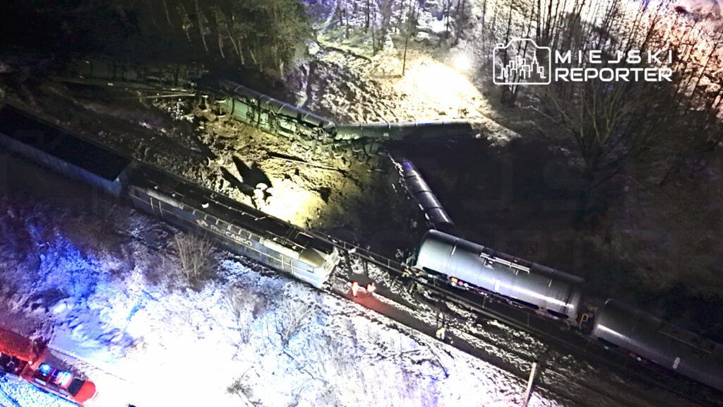 Złomowane wagony kolejowe leżą na boku w zaśnieżonym terenie, a w tle widać pojazdy ratunkowe z włączonymi sygnałami świetlnymi.