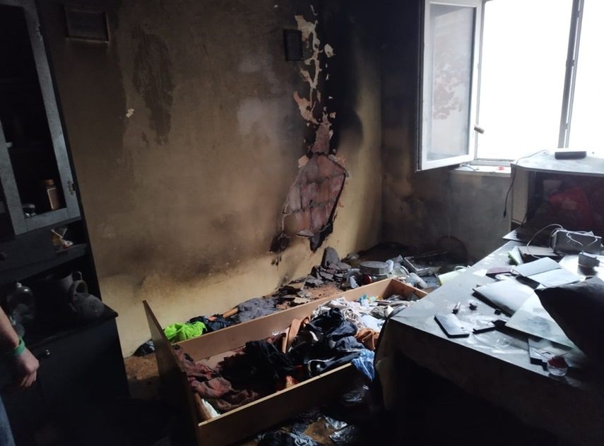Zniszczony pokój z popalonymi ścianami, otwartym oknem i bałaganem na podłodze oraz meblach.