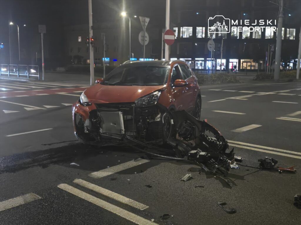 Czerwony samochód osobowy z uszkodzoną przednią częścią stoi na skrzyżowaniu, obok leżą fragmenty karoserii.