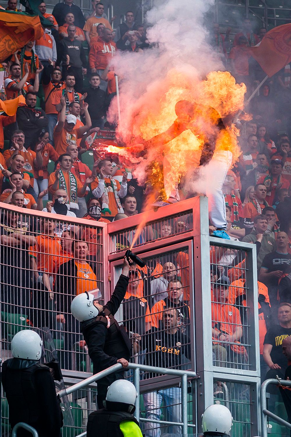 II miejsce, Wydarzenia, zdjęcie pojedyncze. Wrocław. Podczas derbów Śląsk Wrocław – KGHM Zagłębie Lubin na stadionie miejskim kibice odpalali race. Jeden z kibiców Lubina szczególnie się wyróżniał: wchodził na ogrodzenie, machał zapaloną racą w kierunku stewardów. Był zamaskowany. Gdy obok trybun pojawił się szpaler ochroniarzy, jeden miał w ręce coś na kształt gaśnicy, lecz jak się okazało, był to gaz pieprzowy. Gdy go użył, w zetknięciu z racą niespodziewanie powstał pożar i kibic na moment zapłonął jak pochodnia. Nie doznał obrażeń; przed poparzeniem twarzy uchroniła go kominiarka. 26 kwietnia 2014.