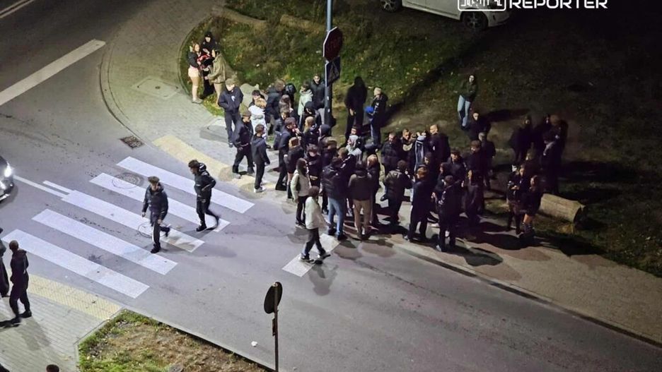 Grupa ludzi w ciemnych ubraniach gromadzi się na przejściu dla pieszych w nocy, obok zaparkowanych samochodów.