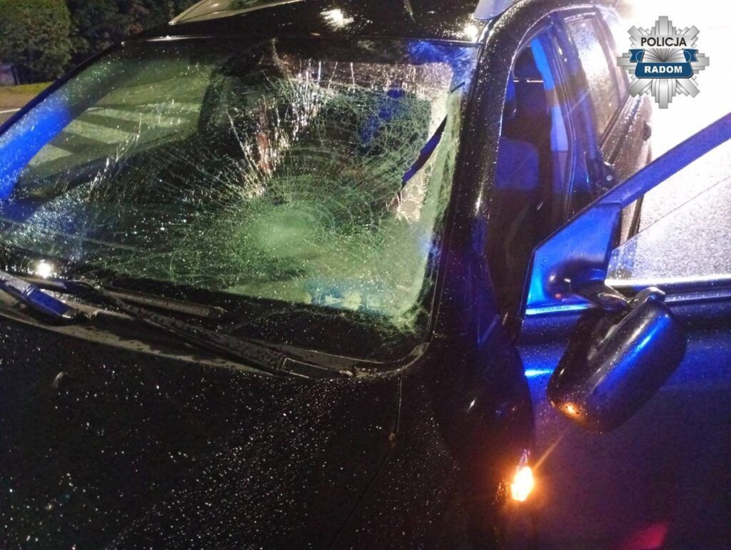 Uszkodzony samochód z rozbitą szybą przednią, zaparkowany w deszczowym otoczeniu nocą. W tle widać niebieskie światła policyjne.