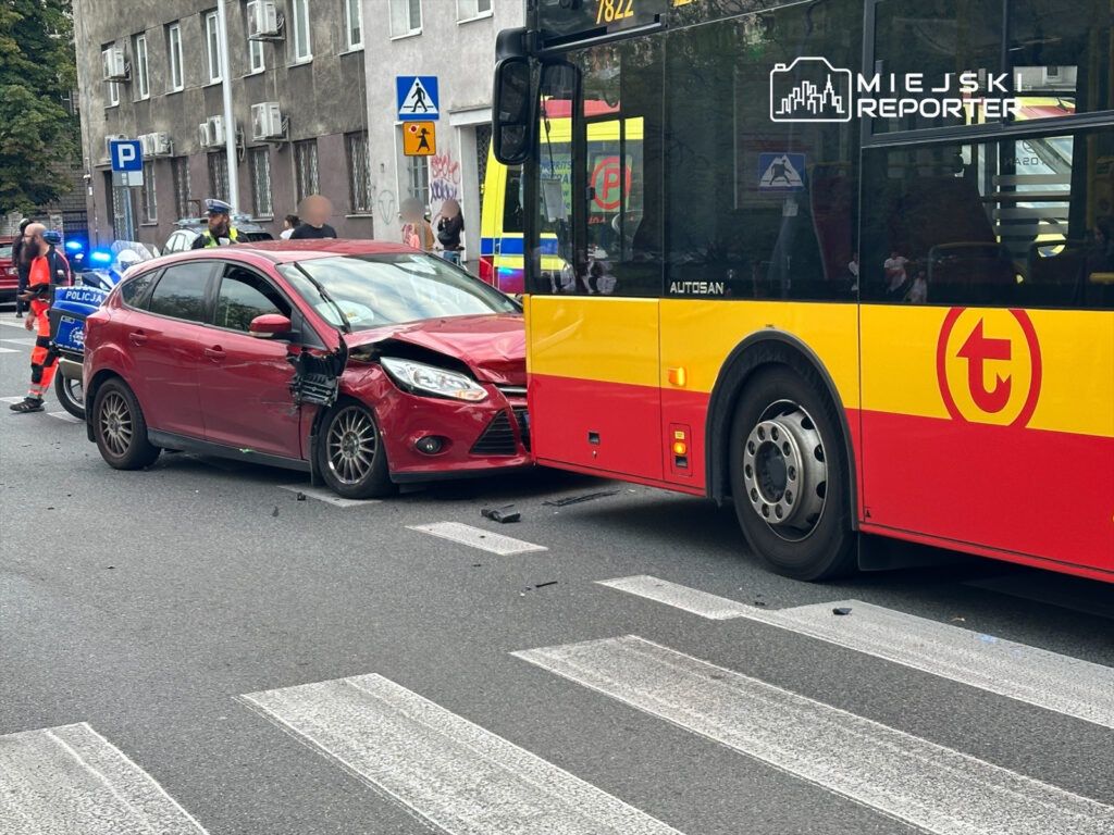 Czerwony samochód osobowy z uszkodzoną przednią częścią zderza się z żółto-czerwonym autobusem miejskim na przejściu dla pieszych. W tle widać funkcjonariuszy policji oraz ratowników medycznych.