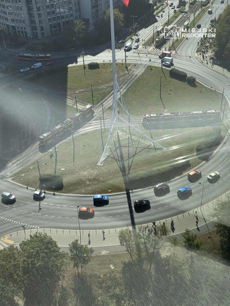 Widok z góry na rondo z tramwajem, samochodami i pieszymi na chodniku, w tle nowoczesne budynki i wysoka flaga.