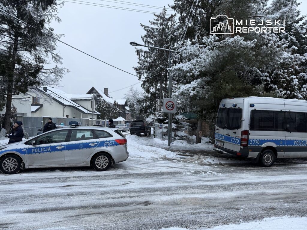 Policjanci rozmawiają obok radiowozu na zaśnieżonej ulicy, w tle widoczne domy oraz znak ograniczenia prędkości.