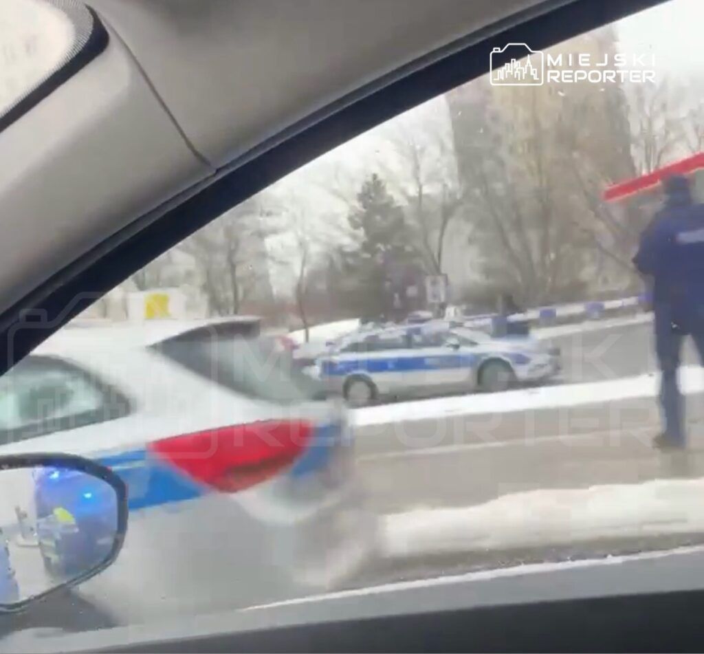 Policyjny radiowóz z włączonymi sygnałami świetlnymi przejeżdża obok grupy funkcjonariuszy na ulicy w zimowej scenerii.