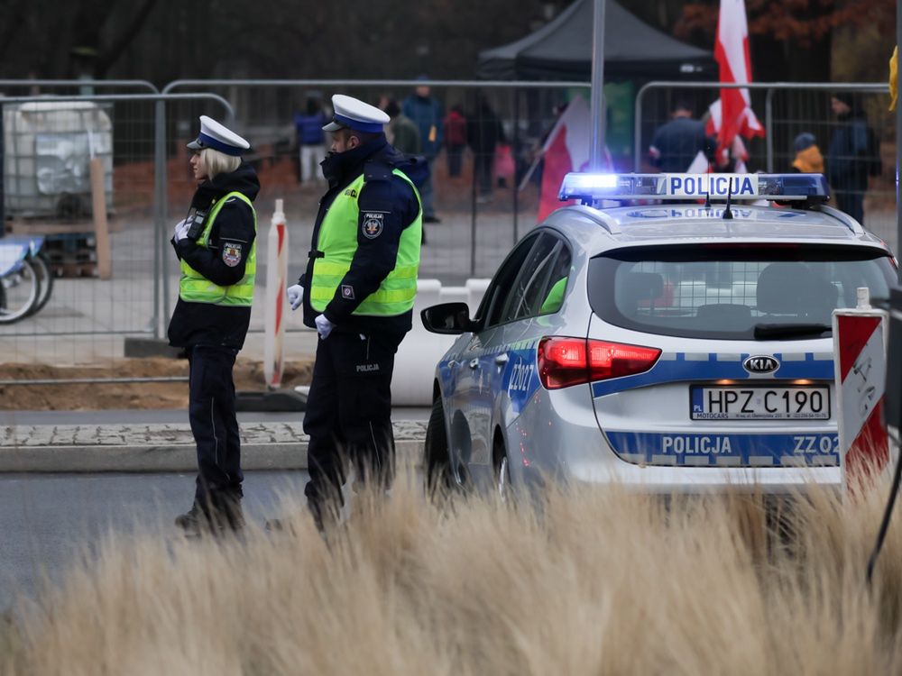 Dwóch policjantów w kamizelkach odblaskowych rozmawia obok radiowozu policyjnego na ulicy.