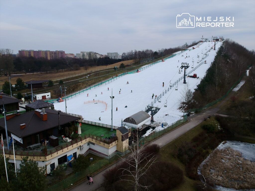 Narciarze zjeżdżają po stoku pokrytym śniegiem, obok widać wyciąg narciarski oraz budynek z tarasem.