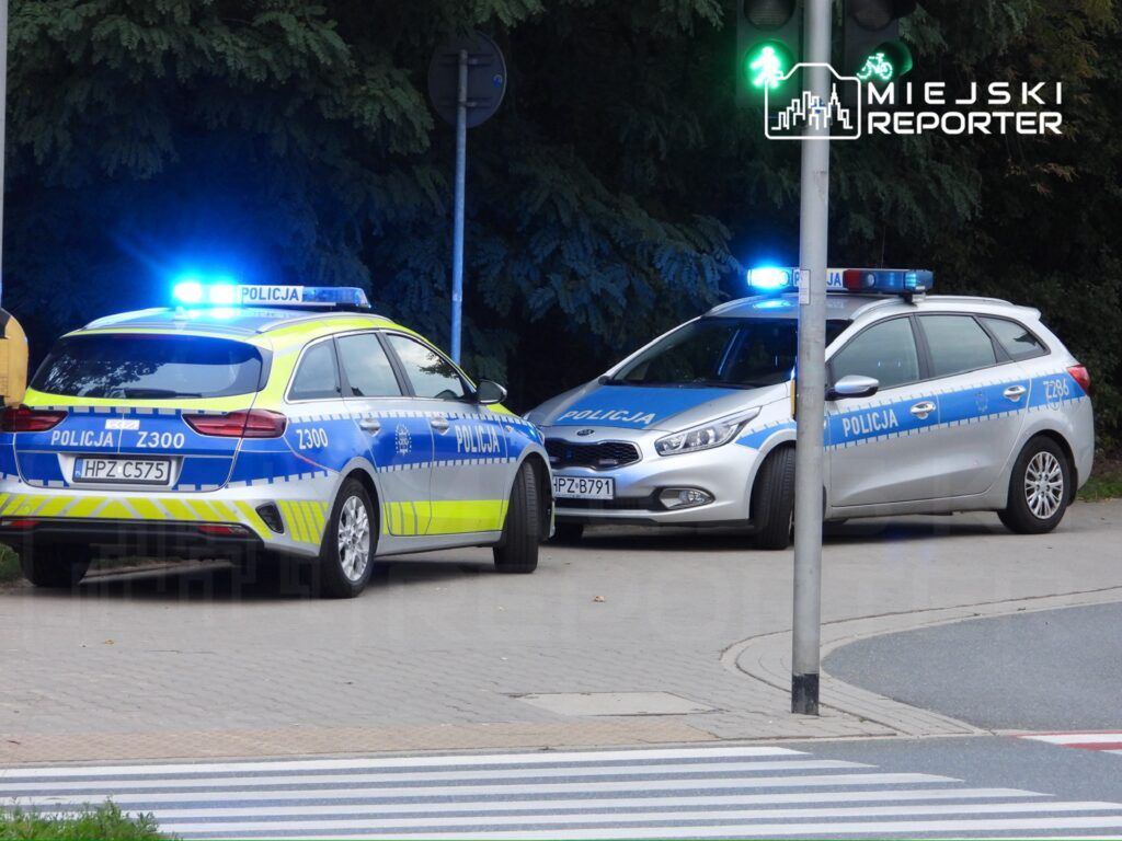 Dwa radiowozy policyjne z włączonymi sygnalizacjami świetlnymi zatrzymane na skrzyżowaniu.