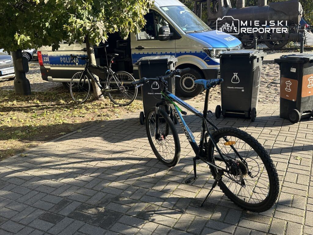 Rower górski zaparkowany obok policyjnego samochodu na ulicy, w tle kontenery na odpady.
