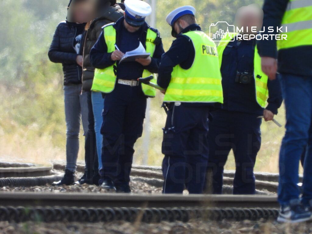 Policjanci w odblaskowych kamizelkach prowadzą rozmowę i spisują notatki na torach kolejowych.