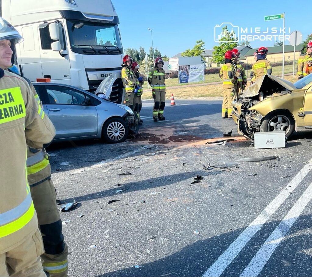 Grupa strażaków w mundurach ocenia skutki zderzenia dwóch samochodów na drodze, wokół widoczne szczątki pojazdów.