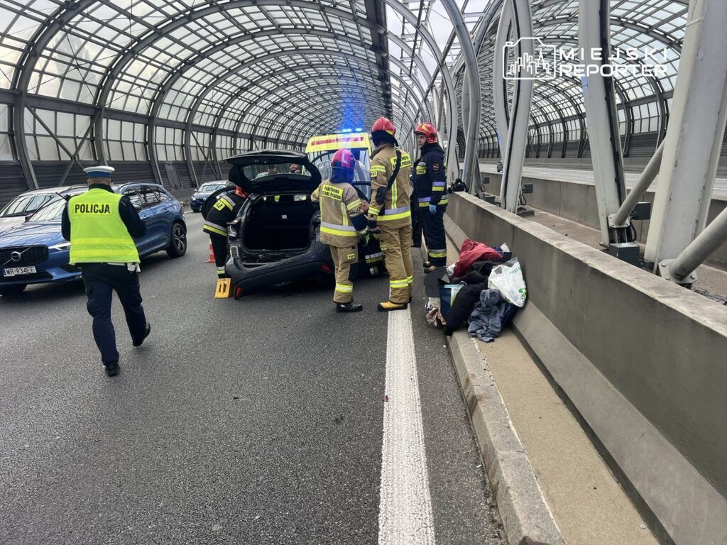 Grupa strażaków i policjantów pracuje przy wypadku drogowym w tunelu, obok leżą ubrania.