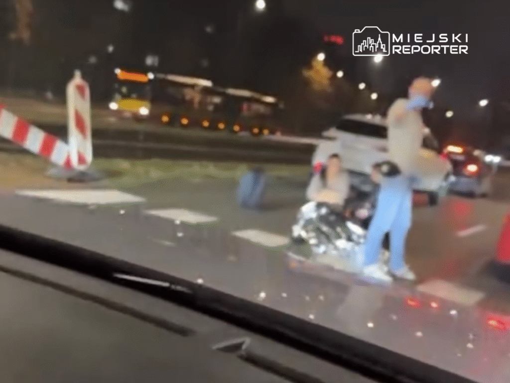 Mężczyzna w niebieskiej koszulce udziela pomocy osobie siedzącej na jezdni, owiniętej w koc termiczny, w nocy.