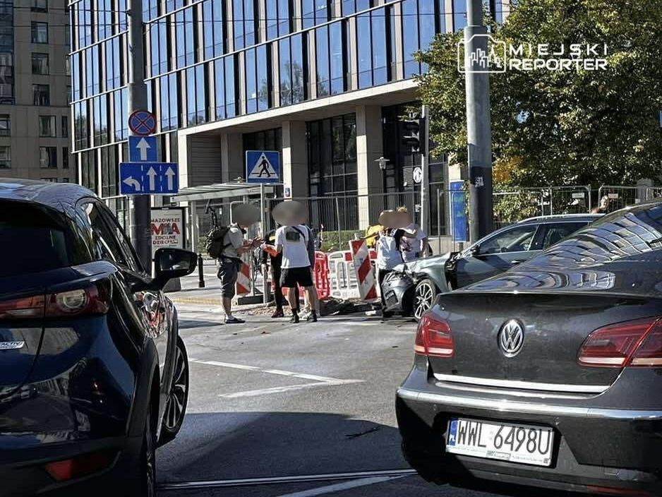 Grupa osób w białych koszulkach rozmawia obok uszkodzonego samochodu na ruchliwej ulicy w mieście.