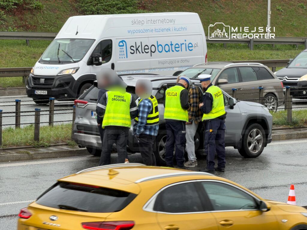 Grupa policjantów i pracowników pomocy drogowej rozmawia z kierowcą przy uszkodzonym samochodzie na ruchliwej ulicy.