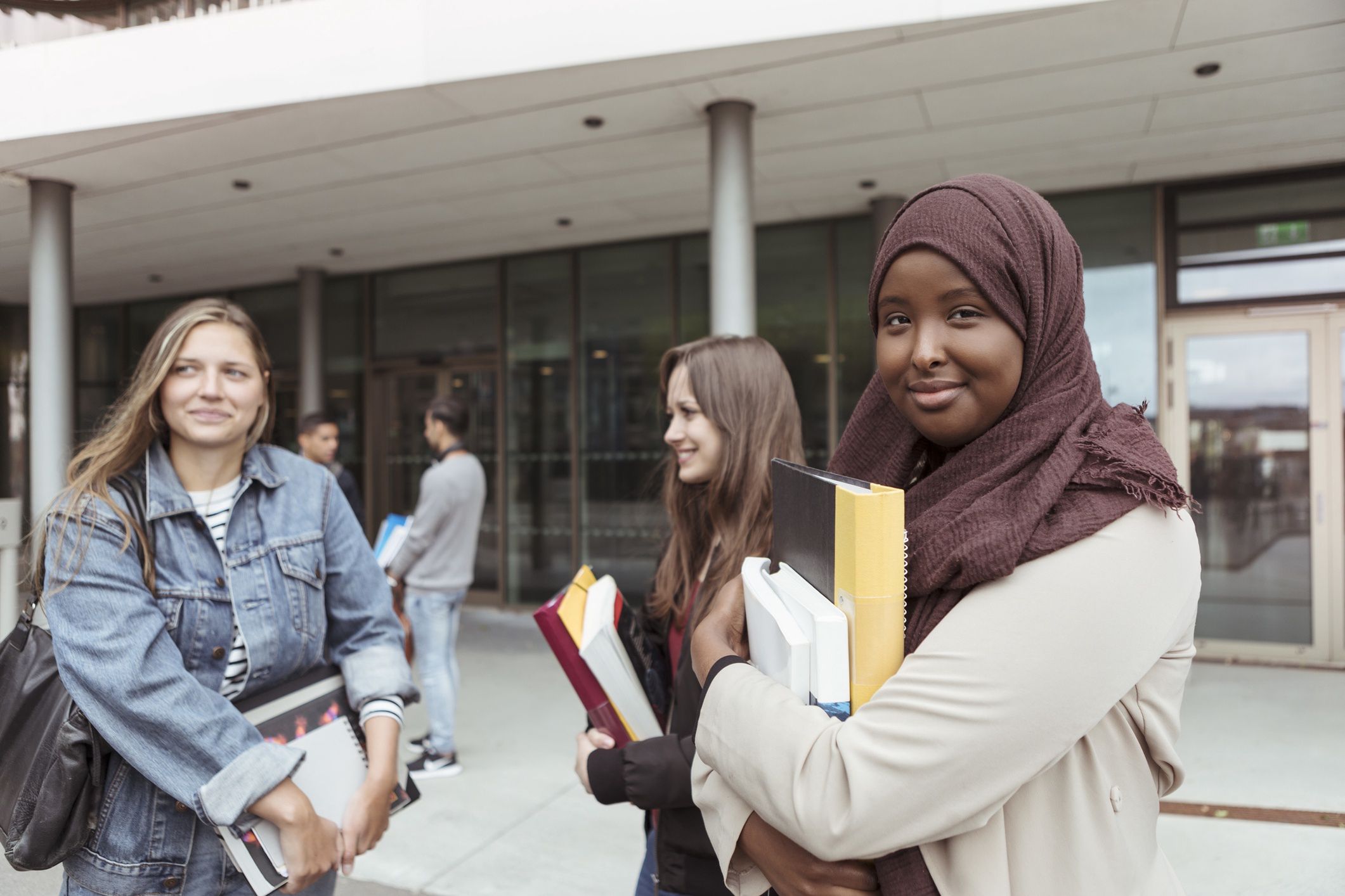 Portrait of student in hijab standing with friends at university campusKentaroo Tryman18-19 years, 20-24 years, african-american ethnicity, arts culture and entertainment, bonding, book, building exterior, campus, casual clothing, color image, communication, day, education, education building, europe, focus on foreground, friendship, high school, hijab, holding, horizontal, incidental people, learning, looking at camera, malmo, millennial generation, islam, multi-ethnic group, outdoors, photography, portrait, post-secondary education, religion, scandinavia, smiling, standing, student, sweden, talking, textbook, three people, togetherness, university, university student, veil, waist up, young adult, young women, exterior