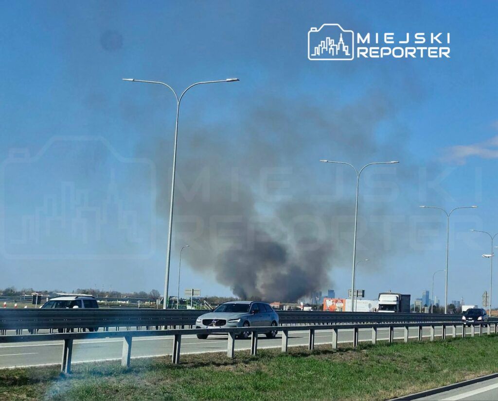 W tle widać gęsty dym unoszący się nad drogą, obok poruszające się samochody na ekspresowej trasie.