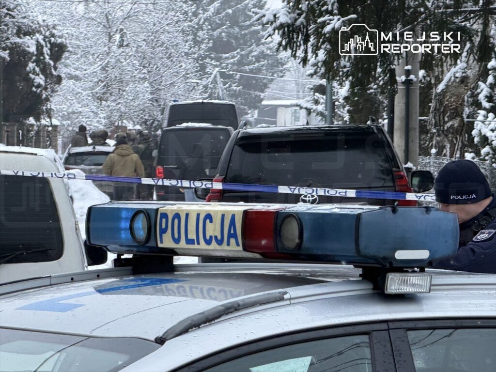 Policyjny radiowóz z włączonymi sygnalizatorami na zaśnieżonej ulicy, w tle grupa funkcjonariuszy i pojazdów.