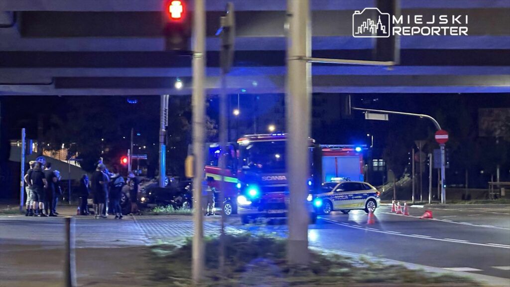 Grupa osób w czarnych ubraniach gromadzi się przy wypadku drogowym, obok interweniujących strażaków i policji.